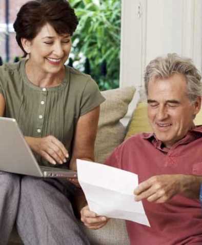 para starszych osób, wyżej siedzi usmiechnięta kobieta z laptopem, poniżej mężczyzna z kartką w ręce, kartka zgięta jak pismo wyjęte z koperty